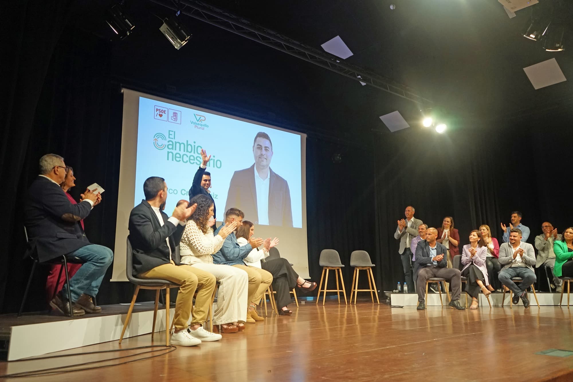 Acto en un teatro: varias personas sentadas en el escenario junto a una pantalla con el lema «El cambio necesario», mientras el público aplaude.