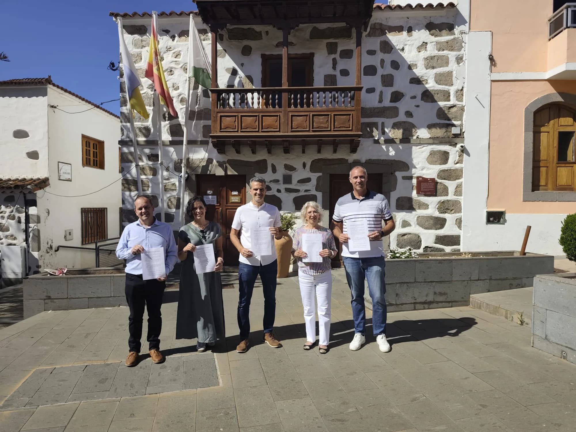 Un grupo de personas posa con documentos en la mano ante el Ayuntamiento de Valsequillo, un edificio de piedra con balcón de madera.